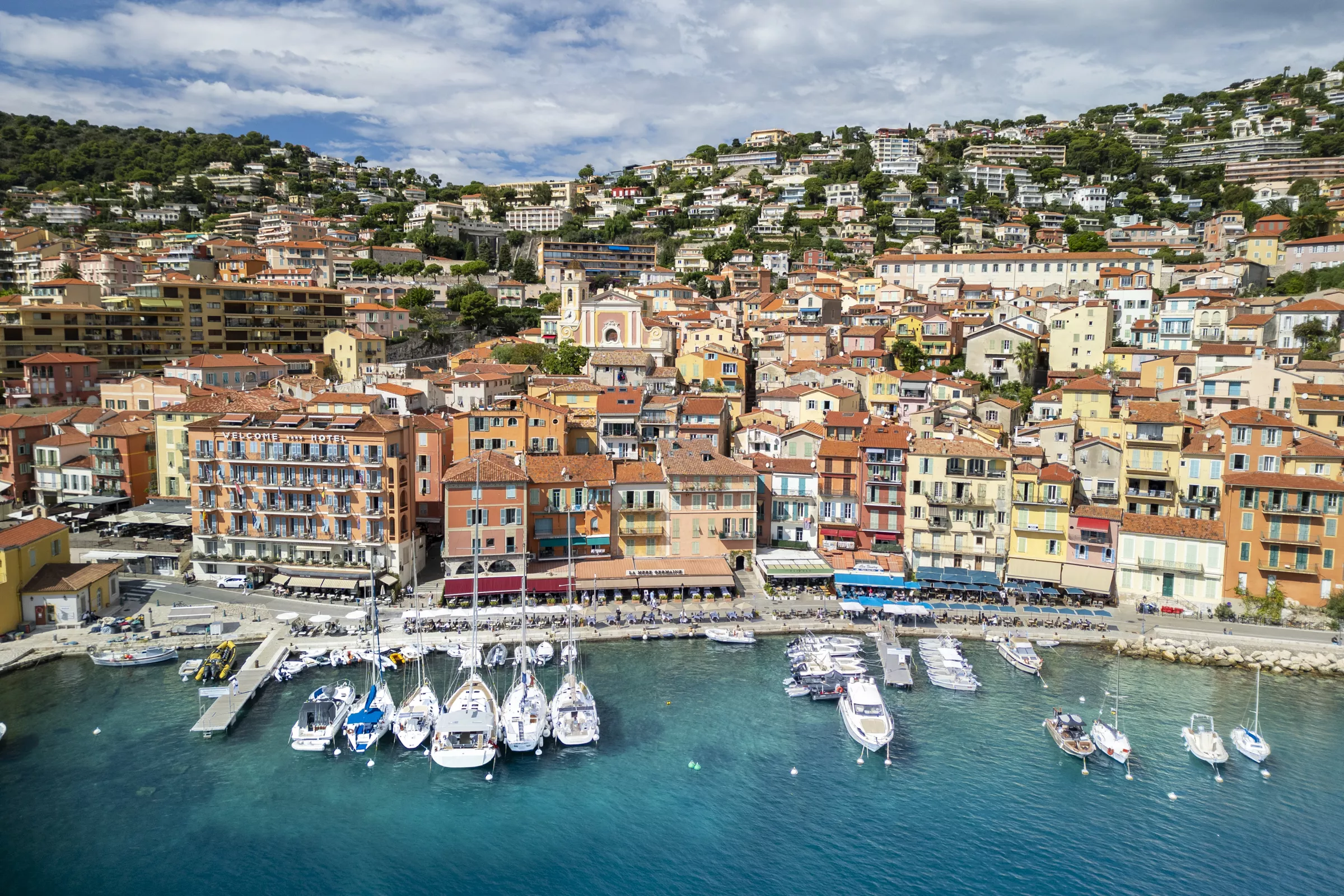 Coastal view of Villefranche-sur-Mer and Beaulieu-sur-Mer with colorful harbor boats, pastel buildings, green hills, and a train running along the seaside near Nice, France.
