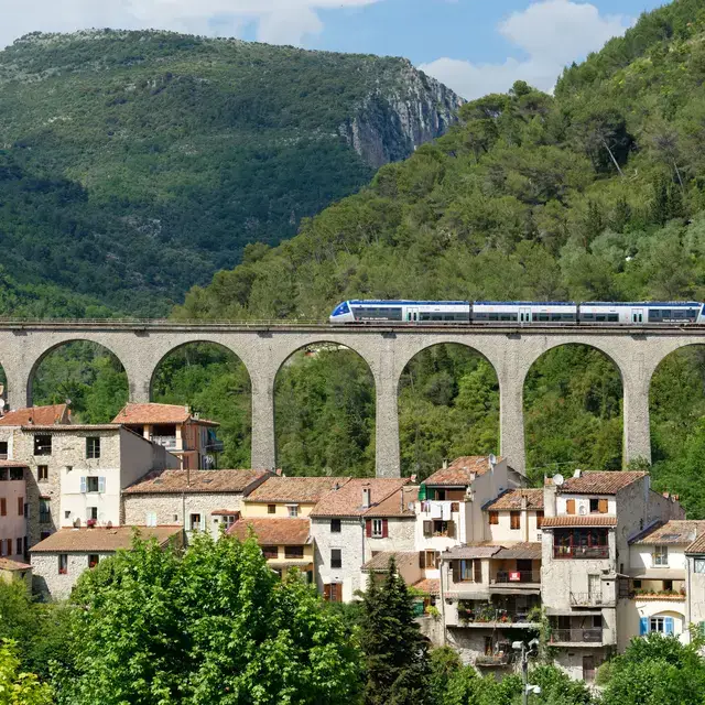 Best Day Trips from Nice by Train, A modern train traveling along a coastal railway near Nice with the Mediterranean Sea and hills with colorful buildings in the background on a sunny day.