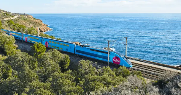 A modern train traveling along coastal tracks with the Mediterranean Sea and colorful cliffs, and the Monaco skyline visible in the background.